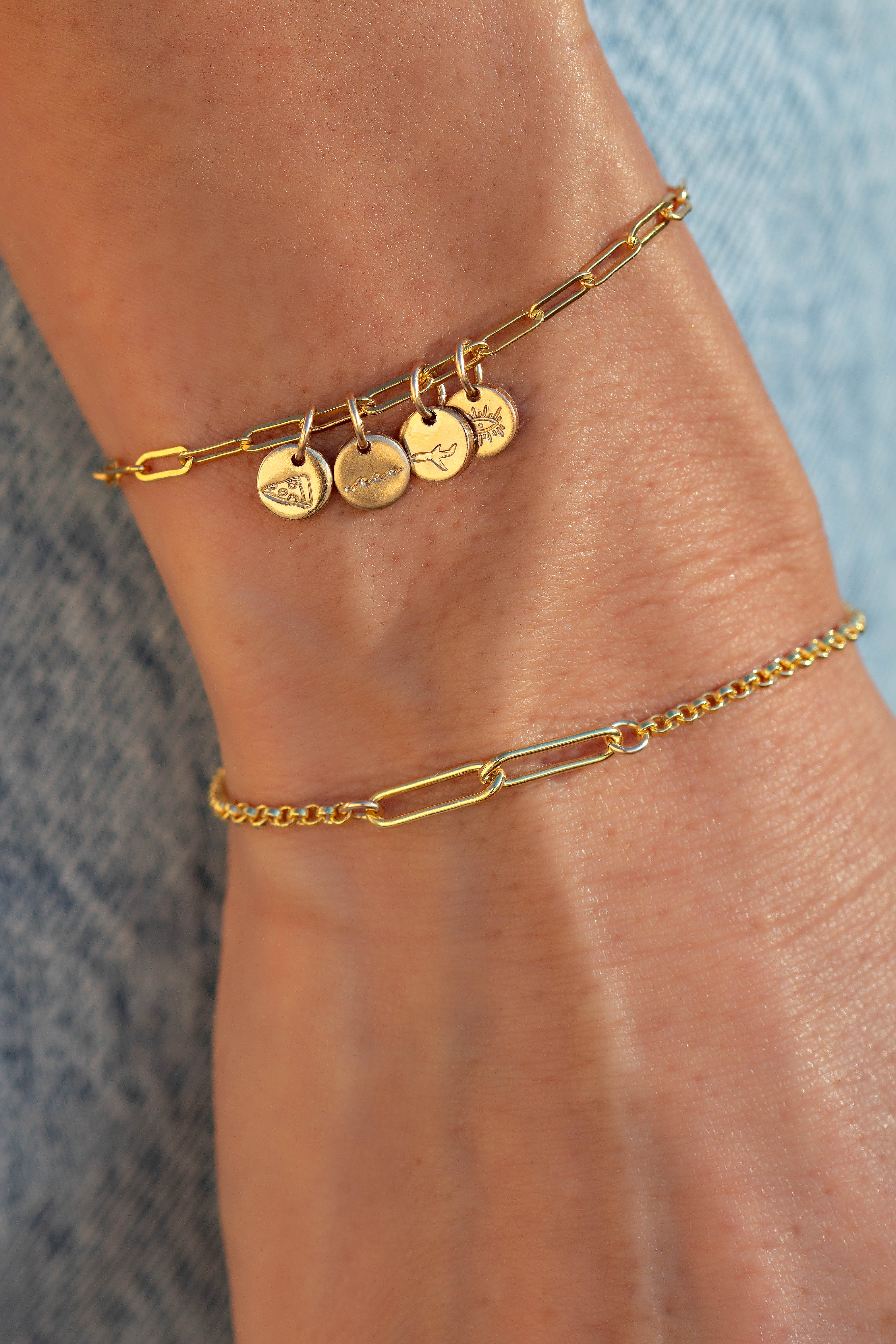 Close-up of a wrist with an interlocking paperclip gold design with multiple round charms on one and the other bracelet a plain interlocking paperclip gold bracelet.