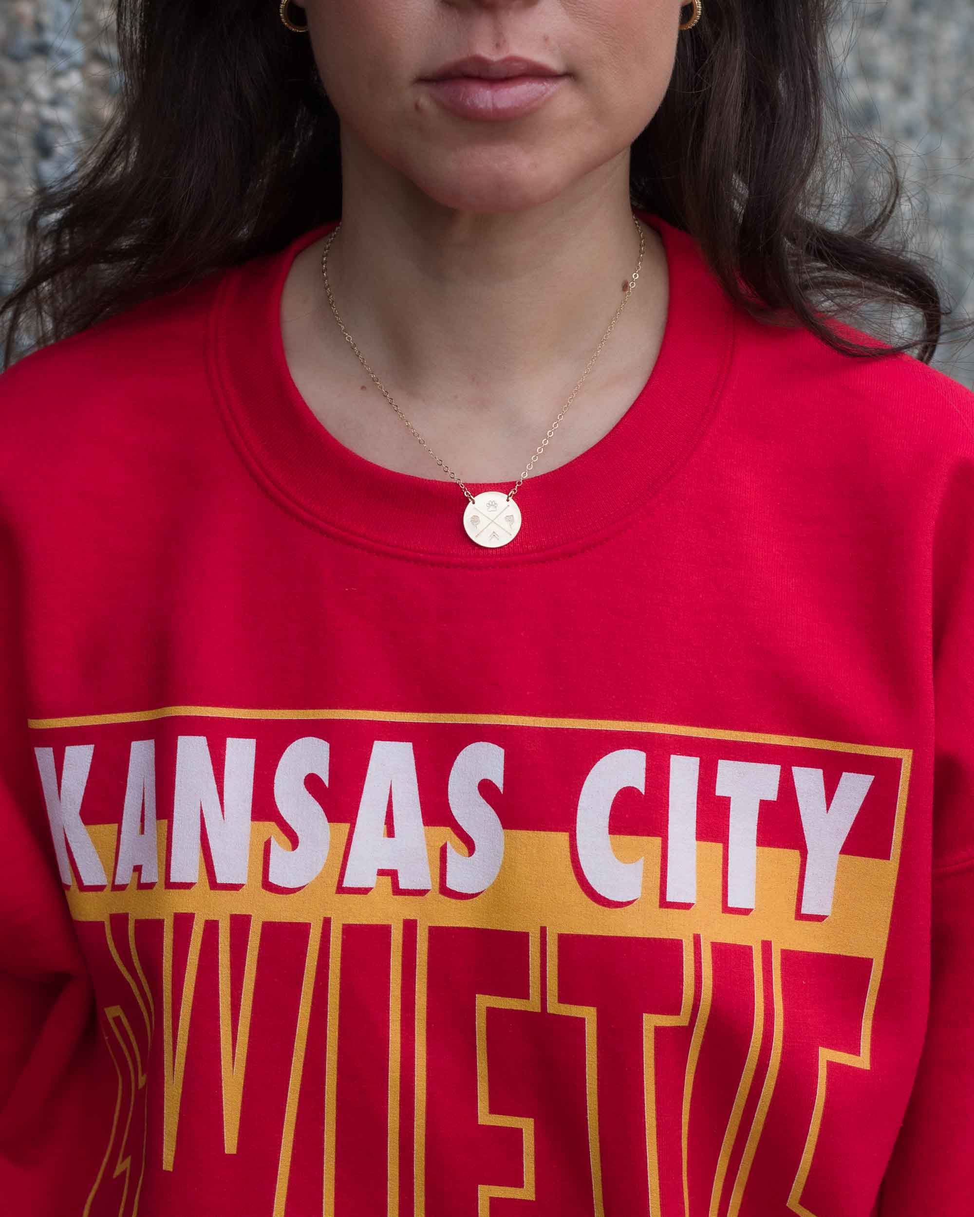 A woman wearing a gold necklace with a round pendant and a gold hoop earrings displayed against a red top as background.