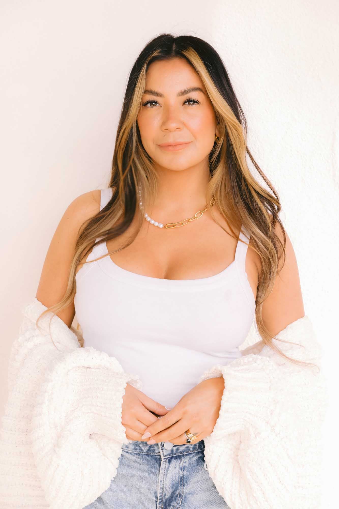 Woman wearing a white tank top with gold and pearl necklace on her neck and a gold ring on her fingers.