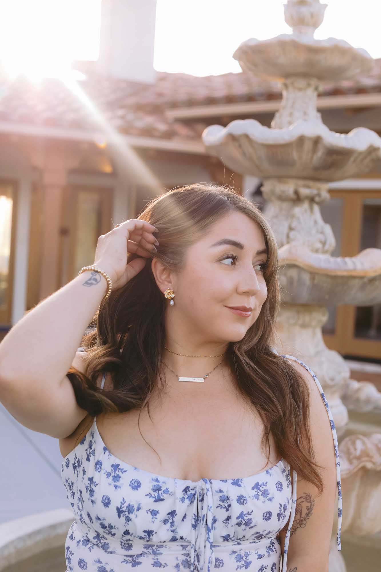Woman wearing a gold earring, gold bracelet, and gold necklace standing in front of water fountain while sunlight shines through