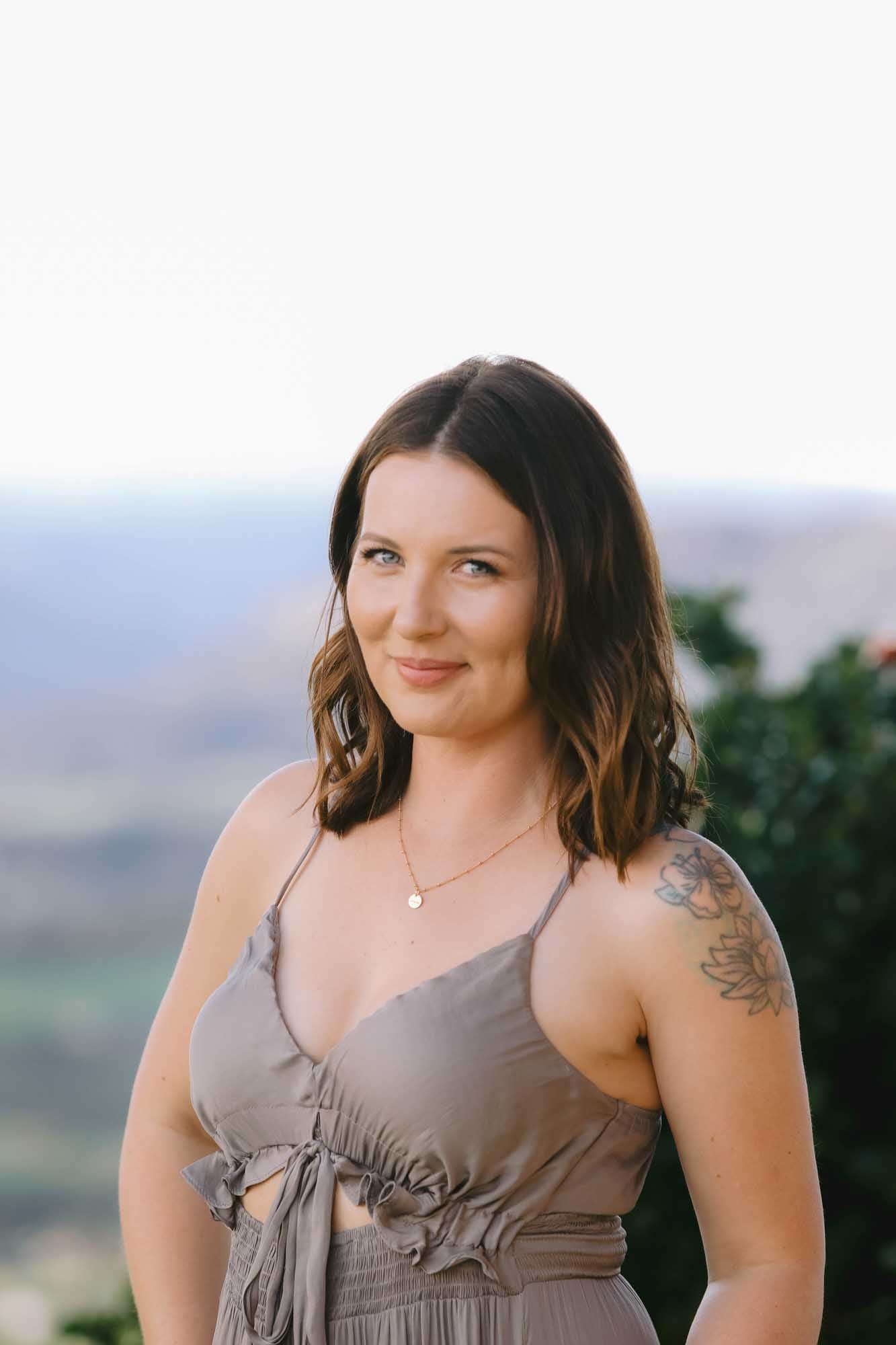 Woman in a sleeveless dress wearing a gold necklace with pendant on an outdoor blurred background.