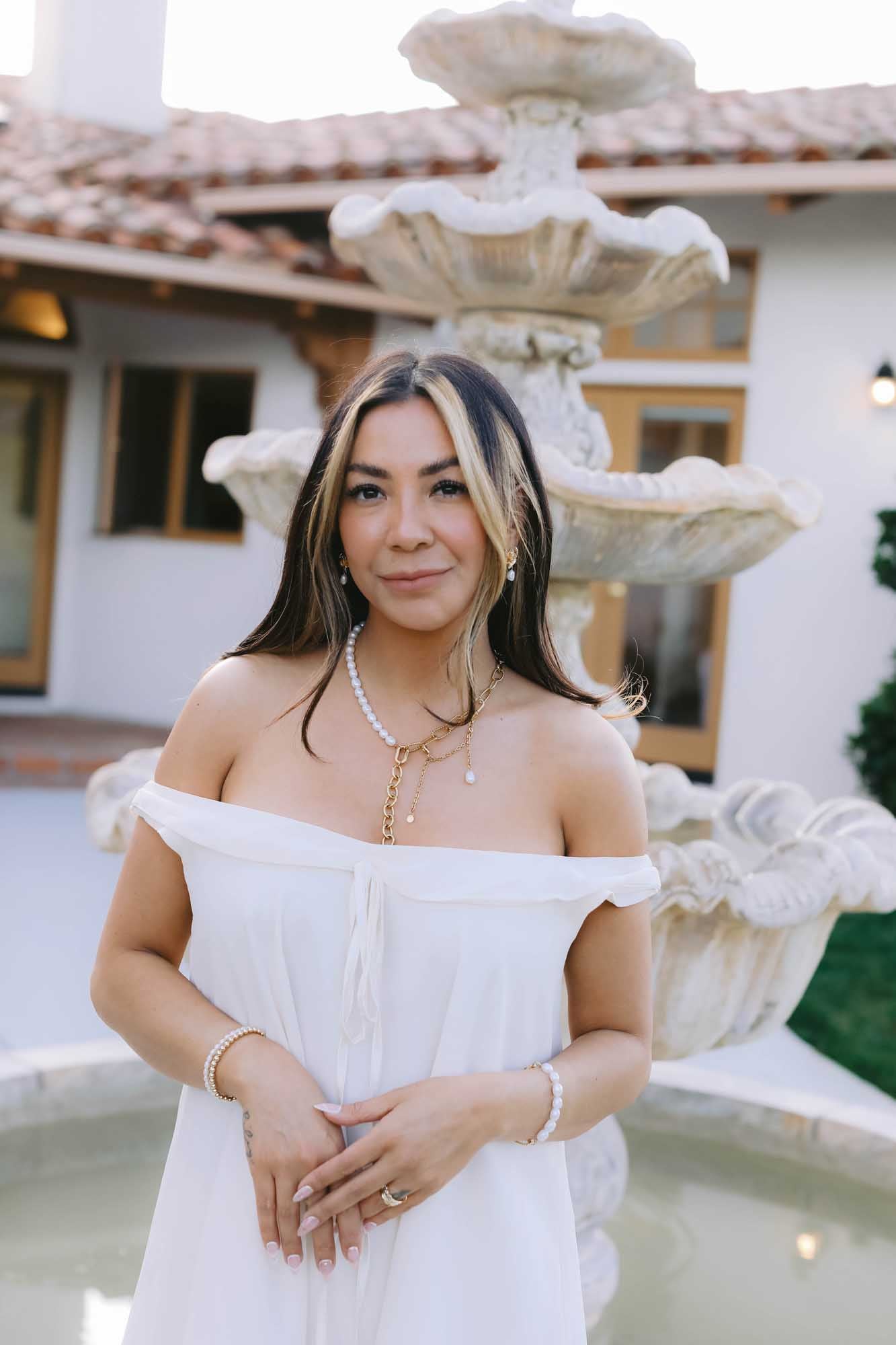 Woman in a white dress standing in front of a fountain wearing gold and pearl necklace, gold and pearl bracelet on each wrist, gold earrings, and gold ring.