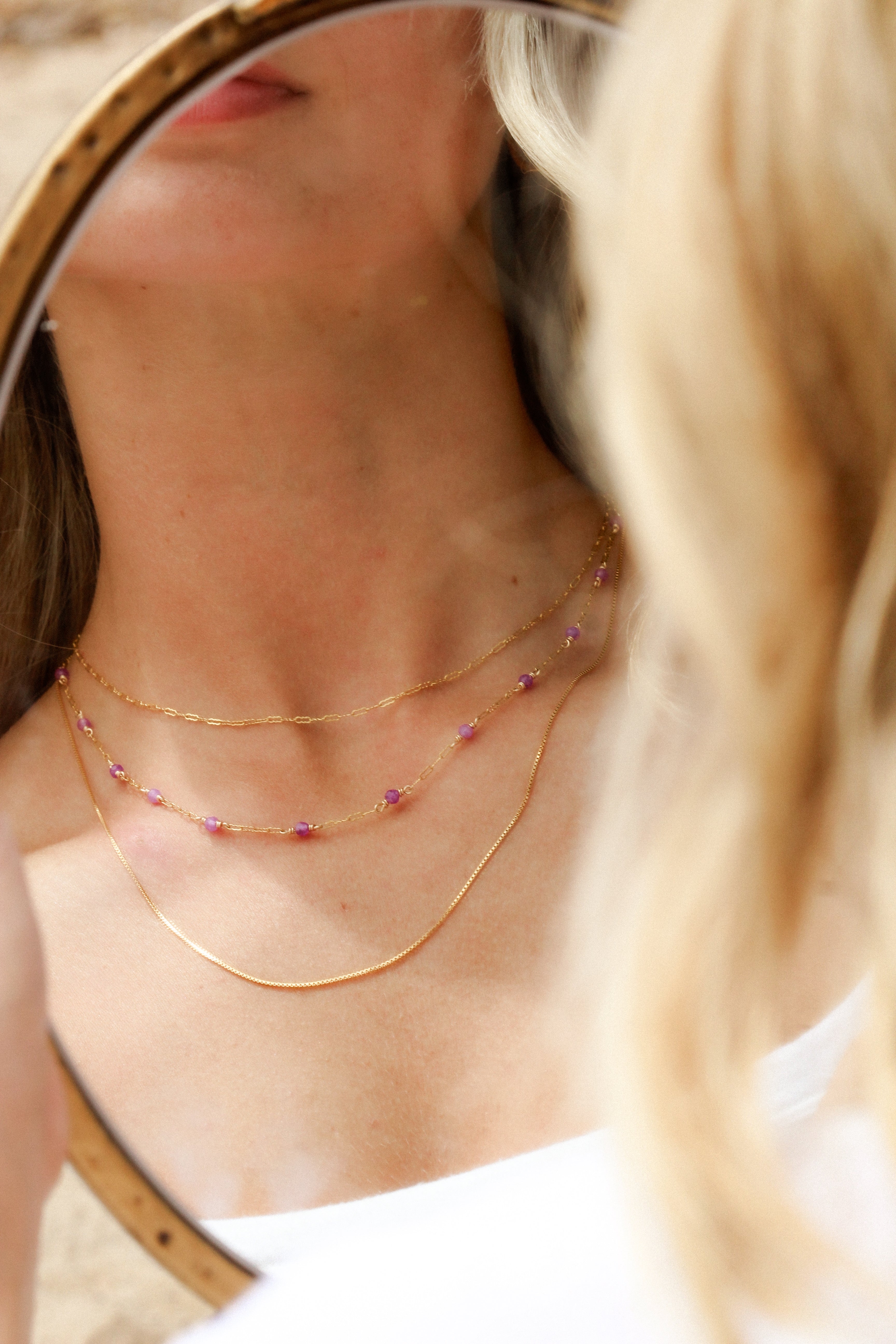 Close-up of a woman wearing multiple gold necklaces including a gold beaded necklace being reflected by a mirror as background.