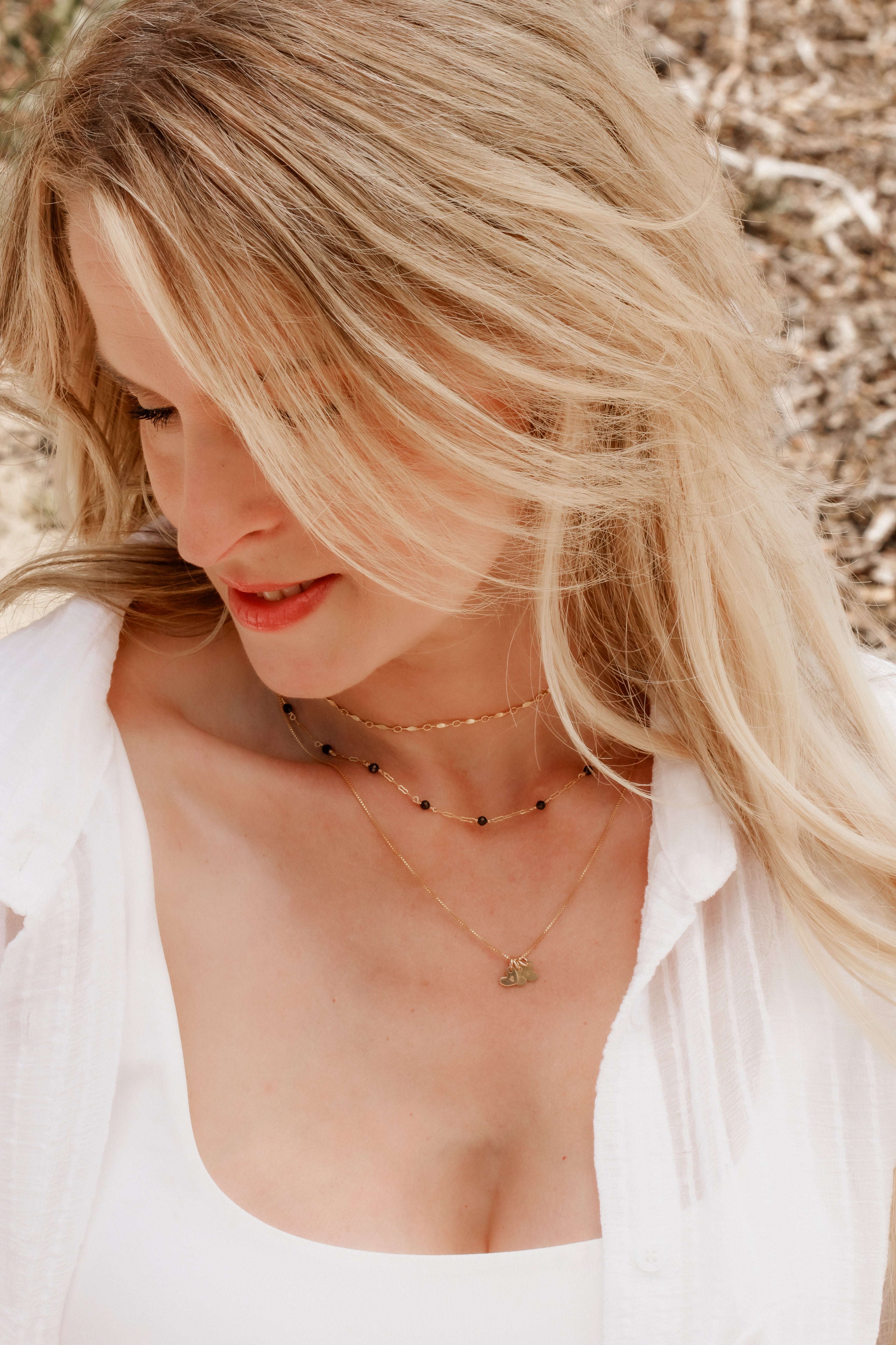 Blonde woman wearing multiple gold necklaces with a blurred natural background.