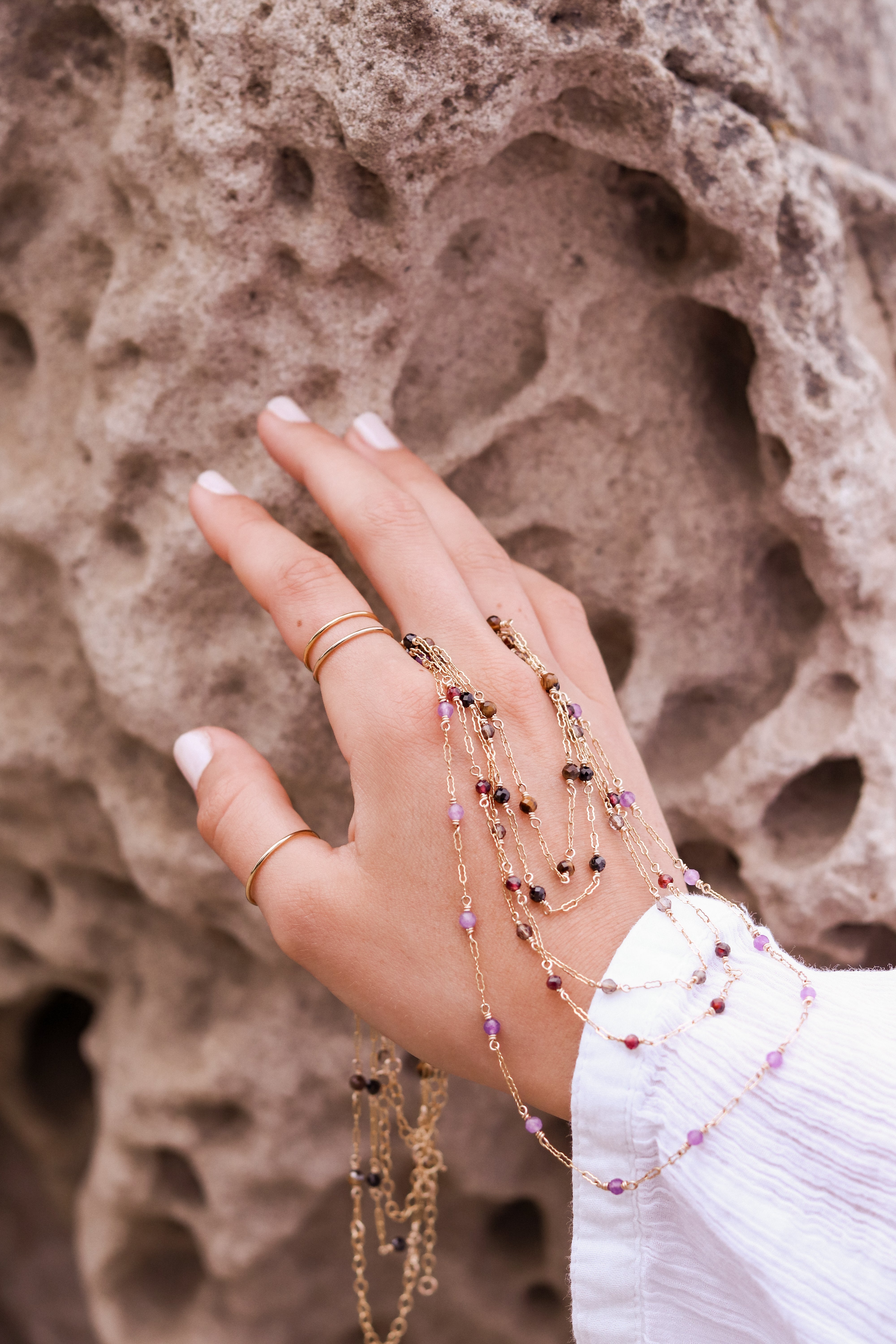 Hand with multiple gold beaded necklaces and gold rings against a textured stone background.