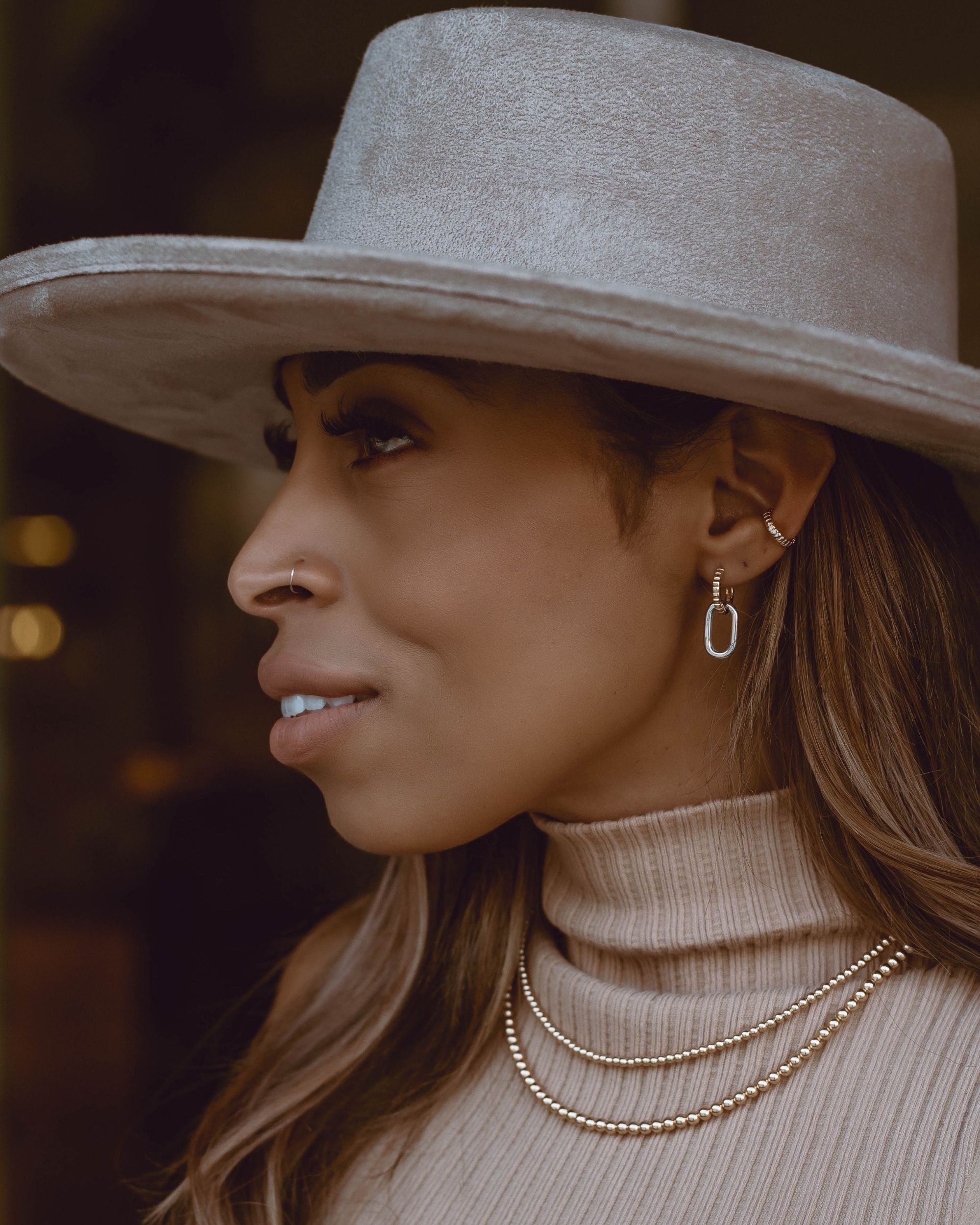A woman wearing two hooped earrings with one contains a paperclip design. She is also wearing a gold ball necklaces on a blurred background.