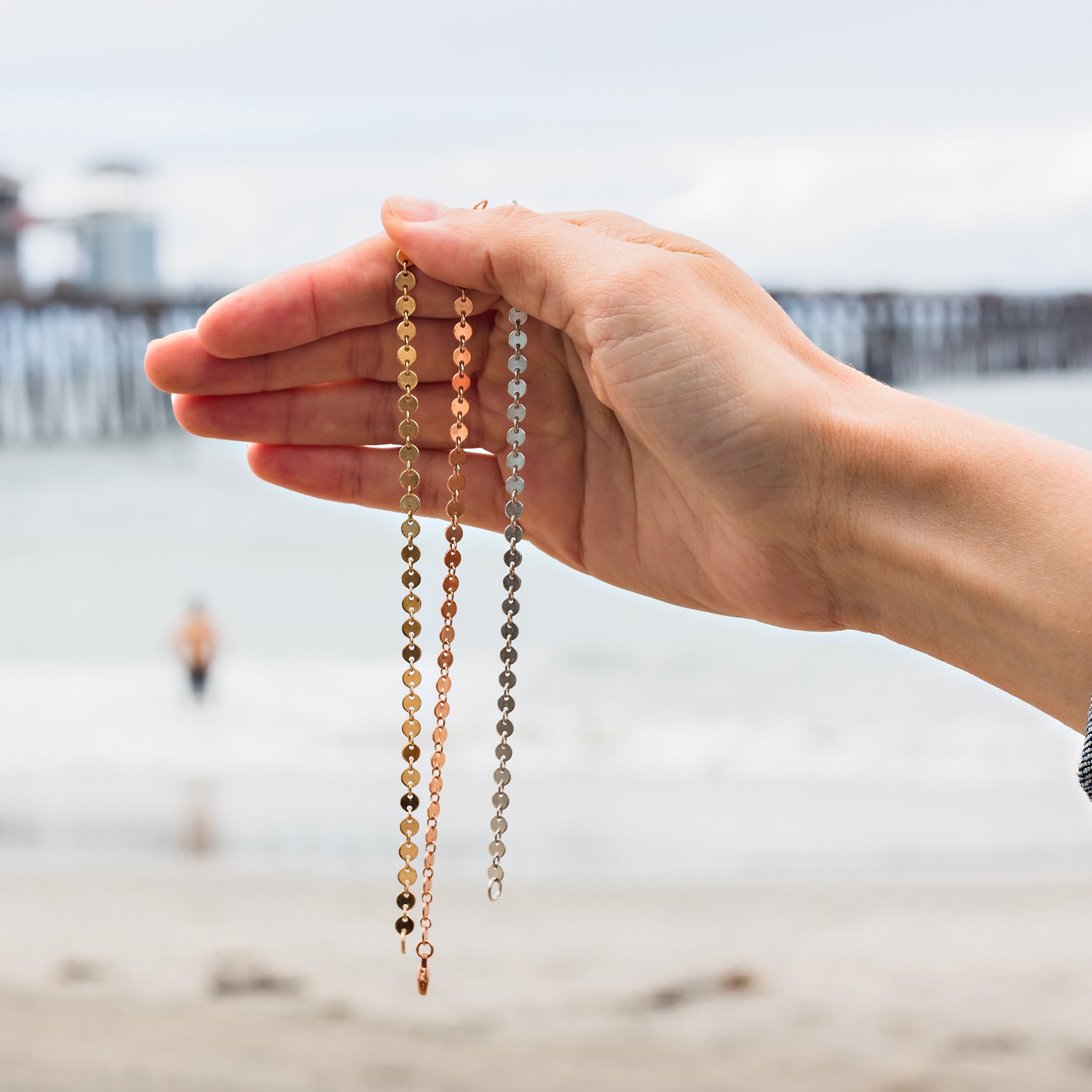A hand holds three delicate disc-link chain necklaces in gold, rose gold, and silver against a blurred beach with a wooden pier and a distant figure