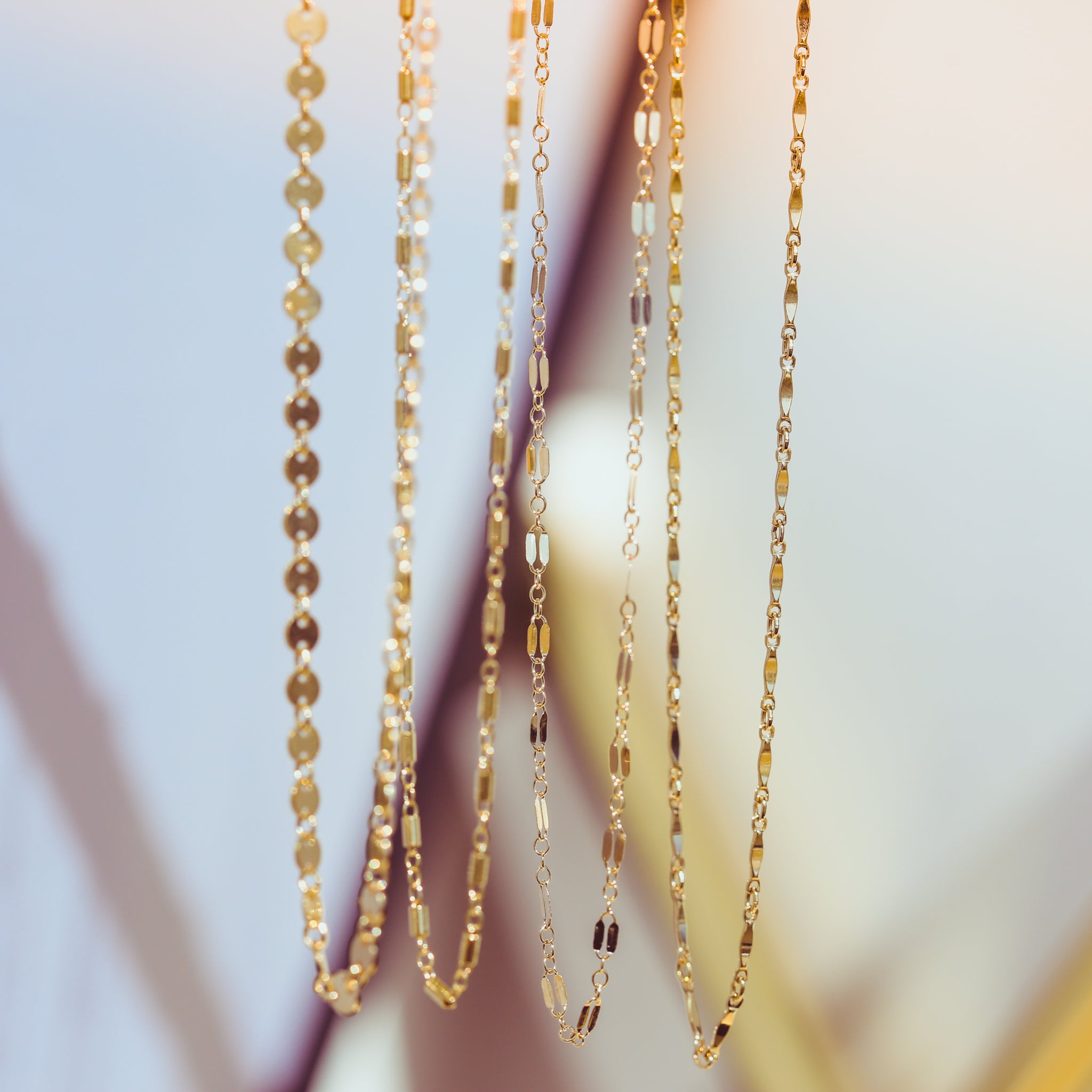 Five elegant gold chains hanging vertically showcasing varied link patterns including flat circular discs and elongated bar shapes shimmering brightly against a soft blurred background