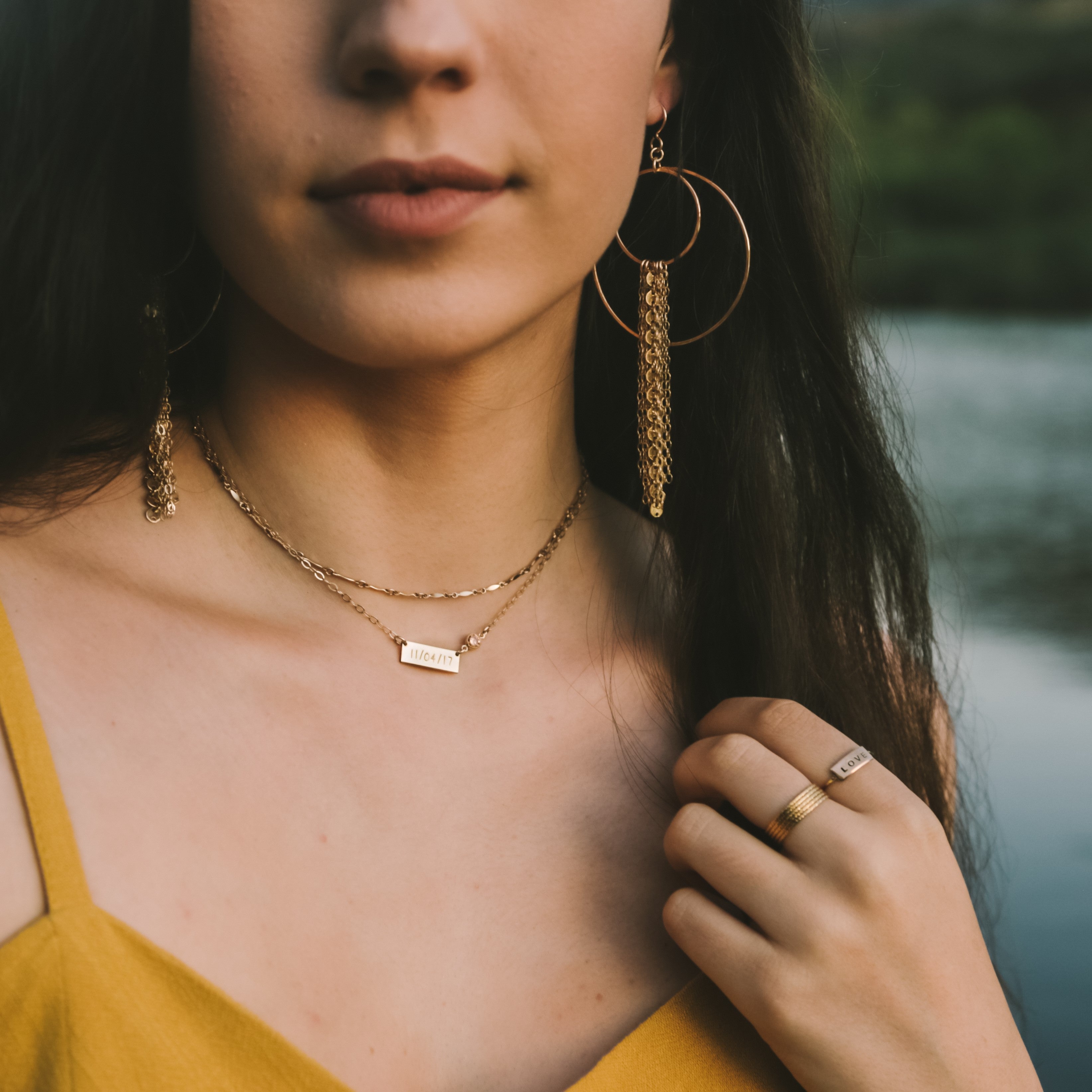 Close-up of a woman featuring large gold hoop earrings with cascading chain tassels, layered gold necklaces with an engraved 11/04/17 bar pendant, and gold rings including one inscribed LOVE, set against a blurry natural water background
