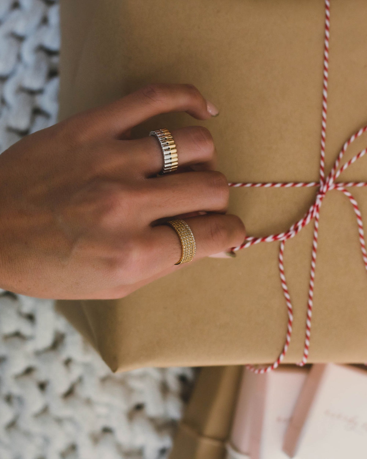 A close-up of a hand wearing a ring with gold and silver design and another gold ring embedded with multiple cubic zirconia surrounding the perimeter of the ring a light brown box in the background.