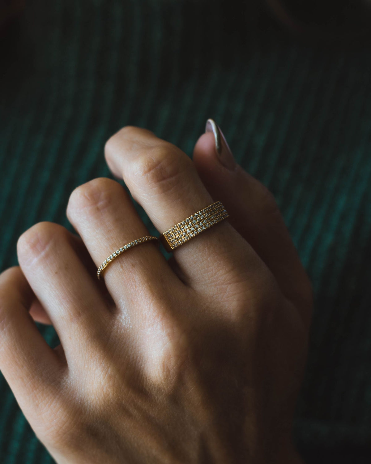 A close-up of a hand with two gold rings with one being thin embedded by cubic zirconia all around the ring. The other gold ring is wider than the previous ring and also decorated with cubic zirconia on a green background.