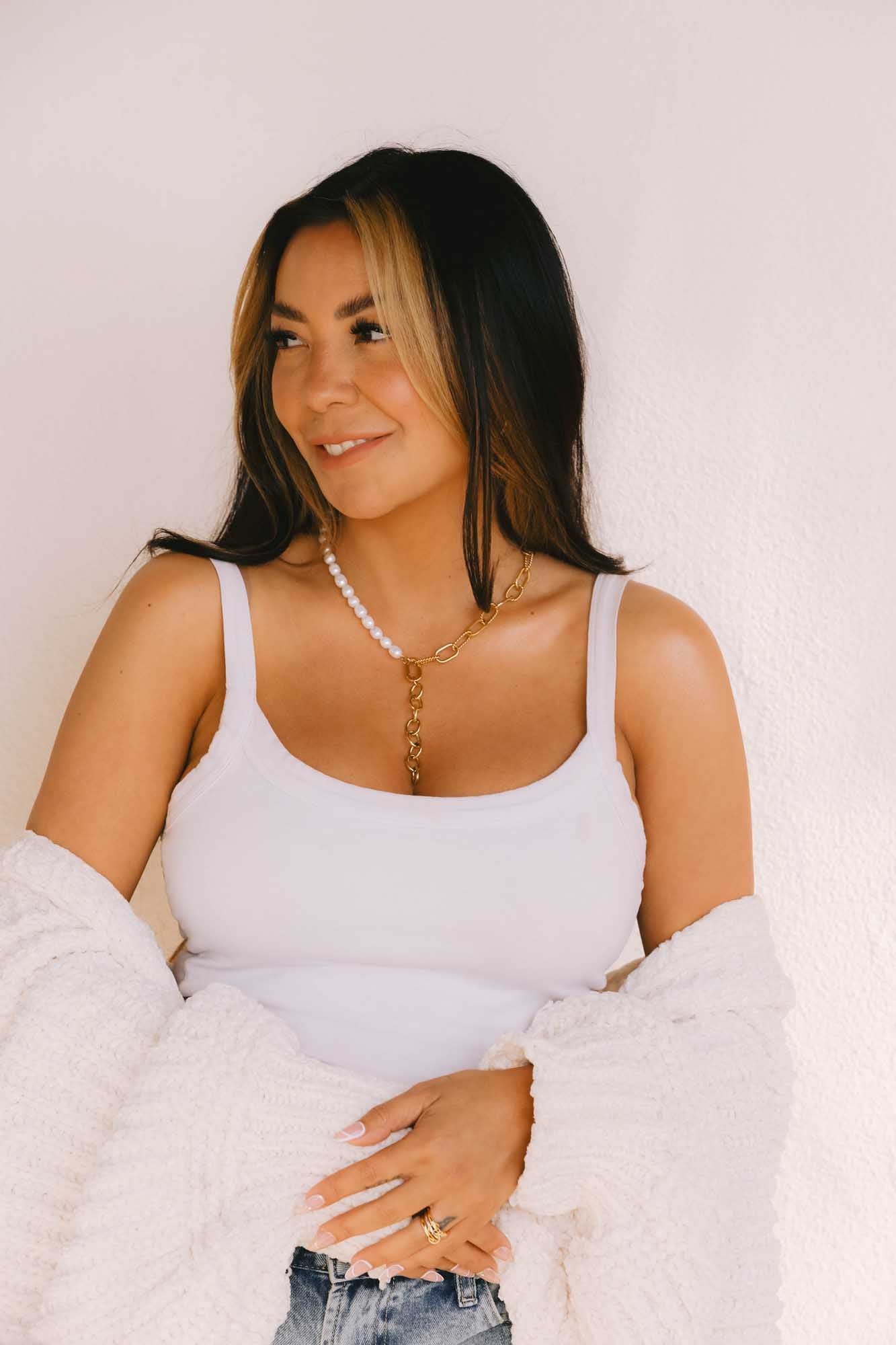 Woman wearing a white tank top and denim with gold and pearl necklace and gold ring.