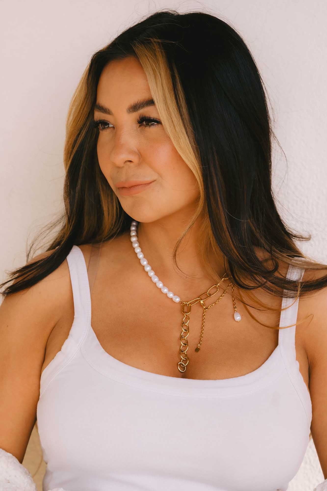 Woman wearing a white tank top and gold and pearl necklace against a plain background
