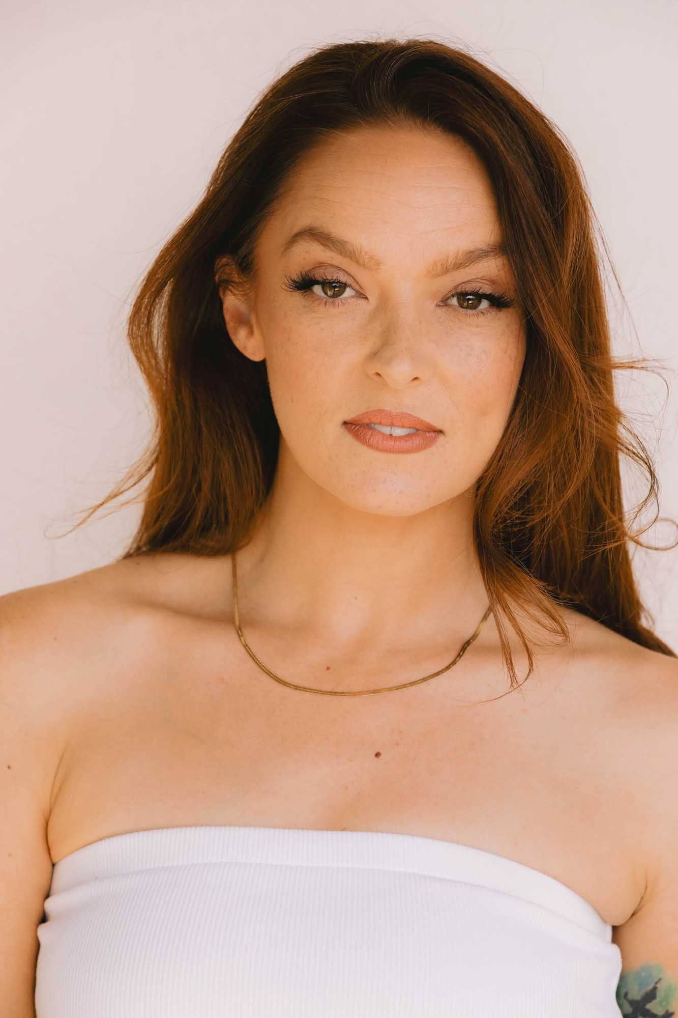 Woman with long red hair wearing a gold necklace on a white background.