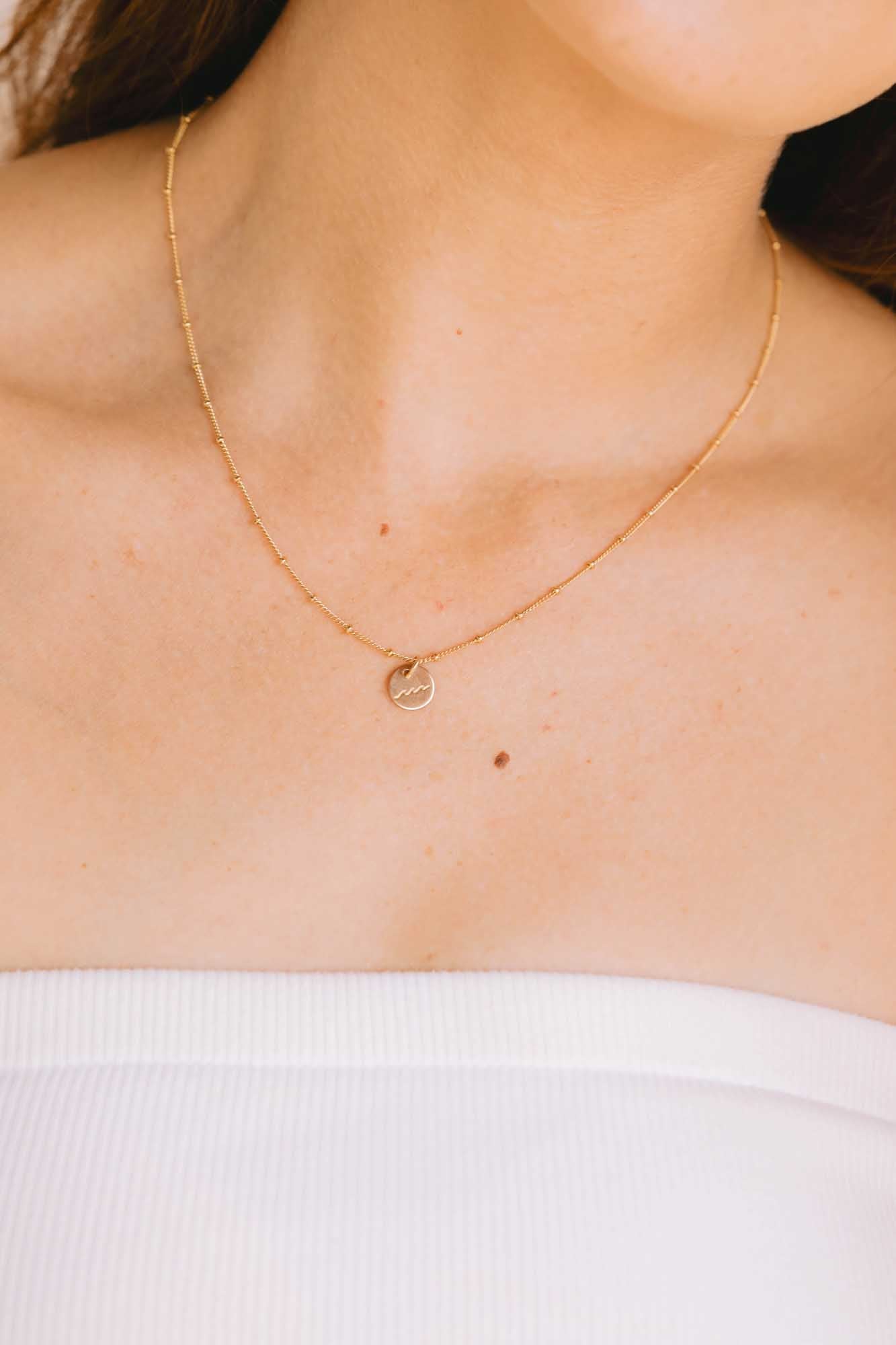 Close-up gold necklace with a round pendant on a person wearing a white top.