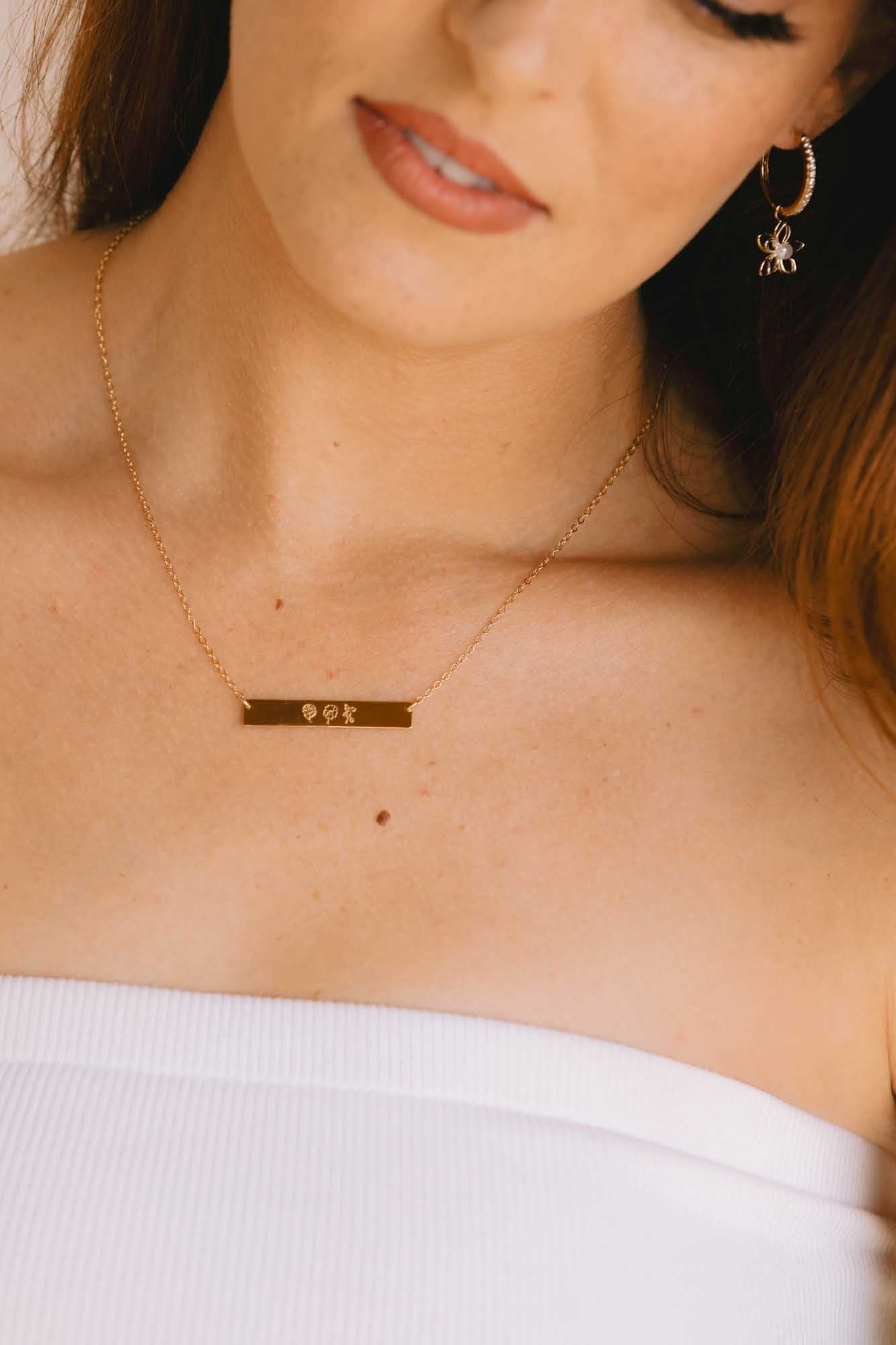 A gold hoop with a star earrings and gold necklace with a bar pendant worn by a person against a neutral background.