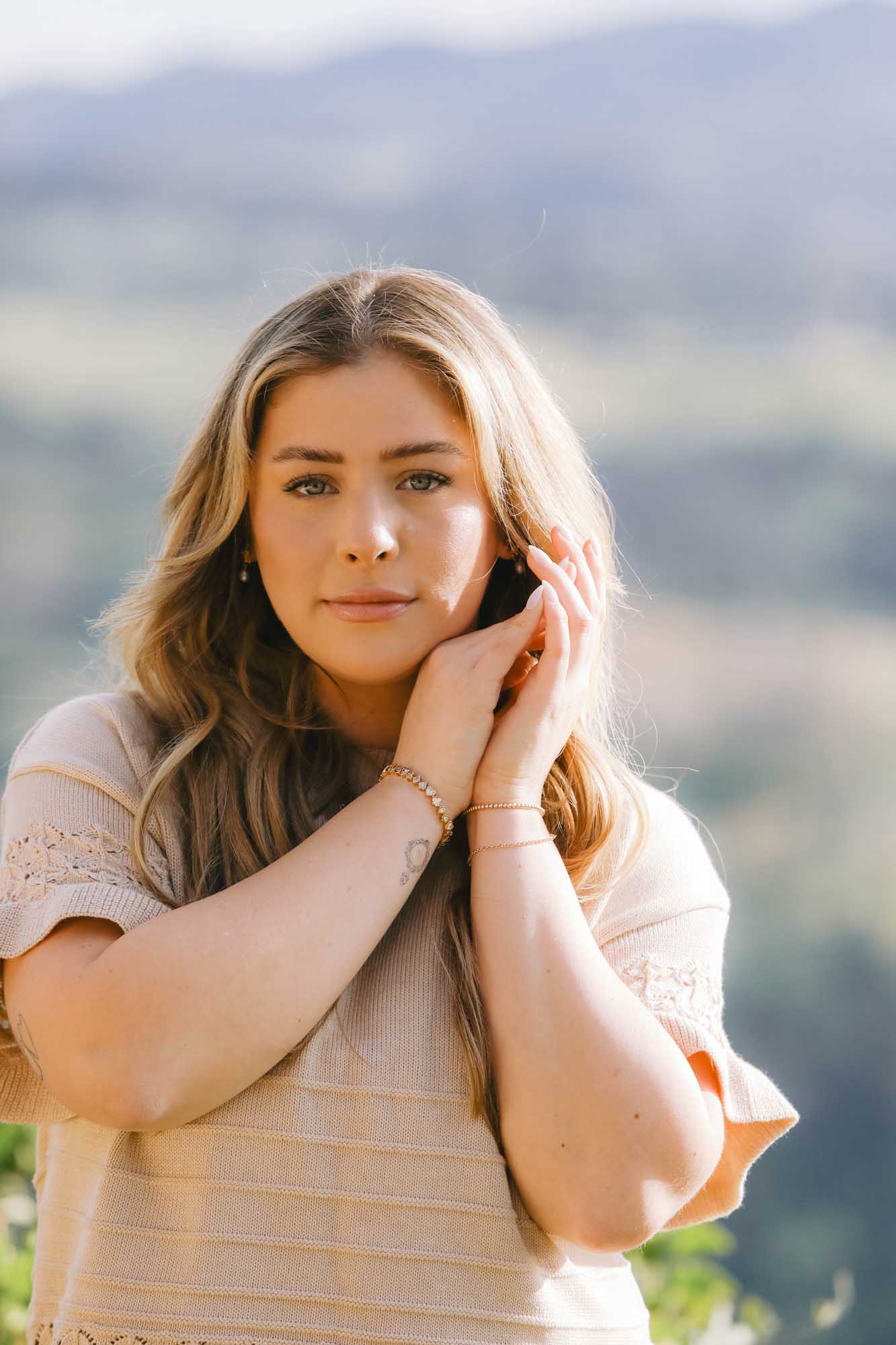 Woman with hands near face wearing gold bracelet and earrings standing outdoors with blurred natural background