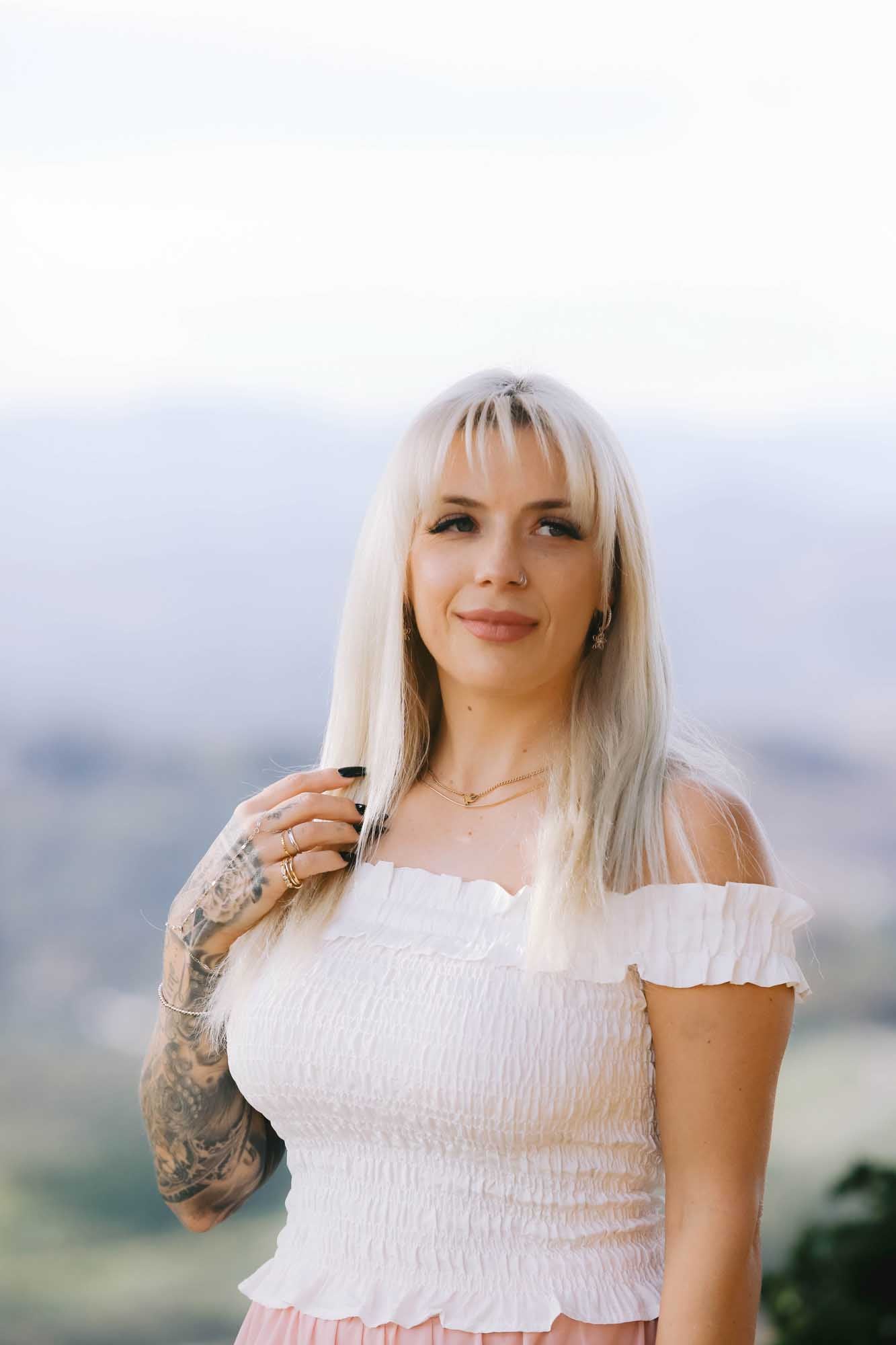 Woman posing while wearing multiple rings, a bracelet, earrings, and gold necklace on a blurred background