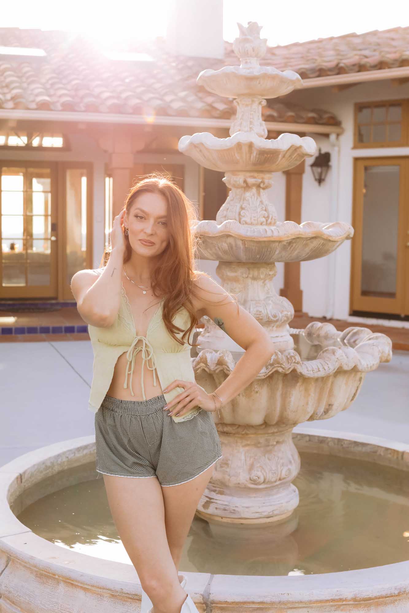 Woman posing by a fountain in a sunlit outdoor setting wearing a gold necklace.
