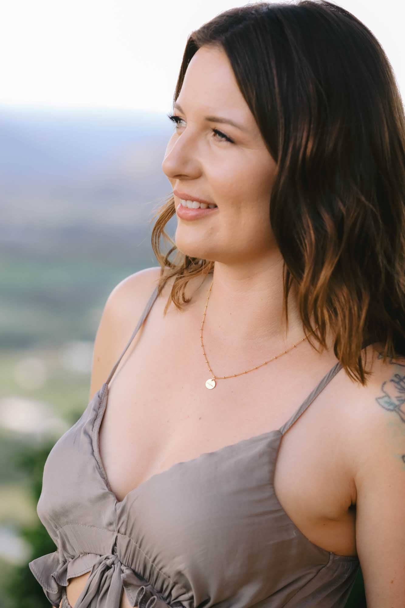 Woman wearing a gold necklace with pendant on a blurred outdoor background.