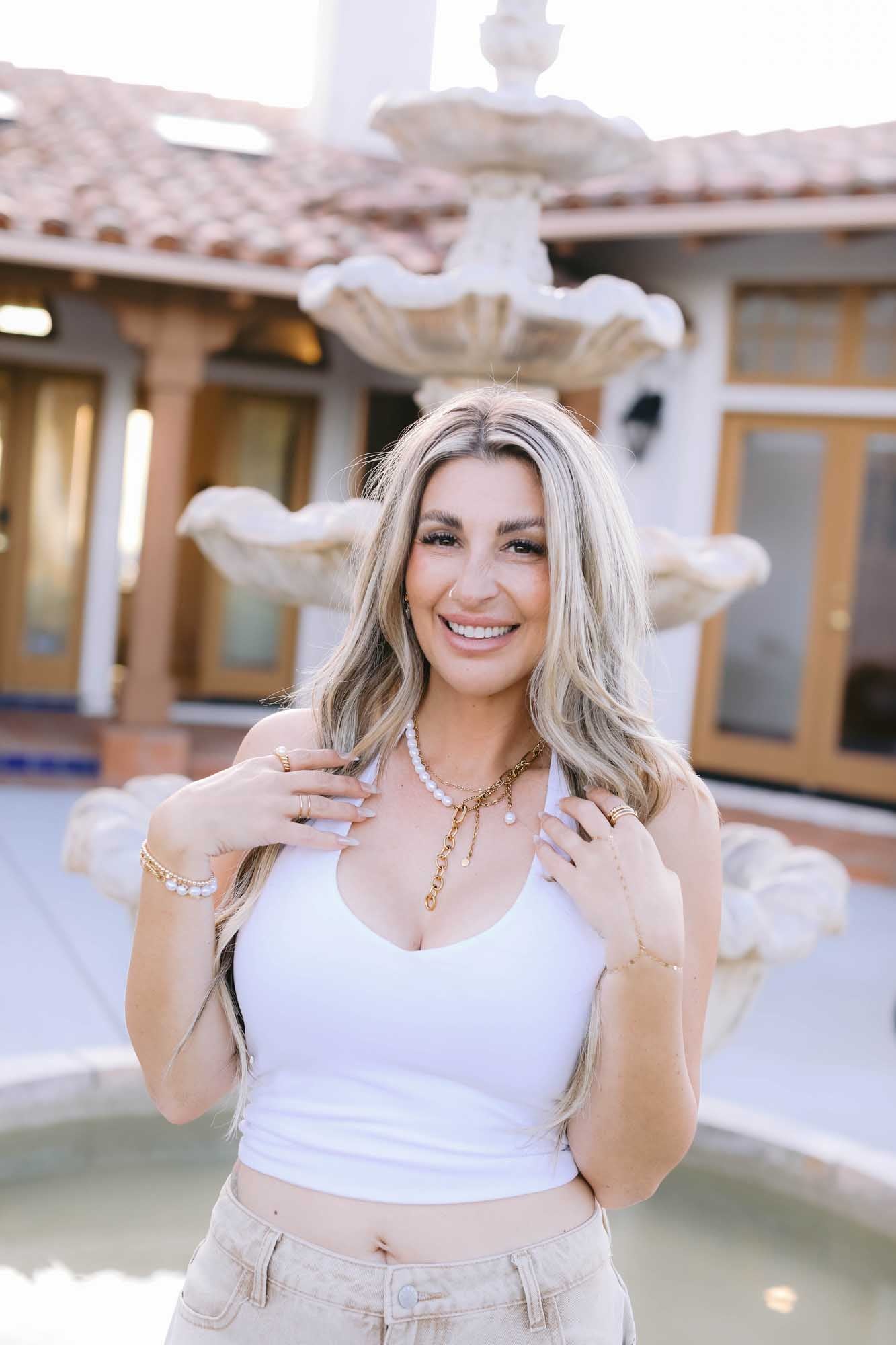 Woman in a white tank top wearing a gold and pearl necklace, gold and pearl bracelet on right wrist, and gold bracelet on left wrist.