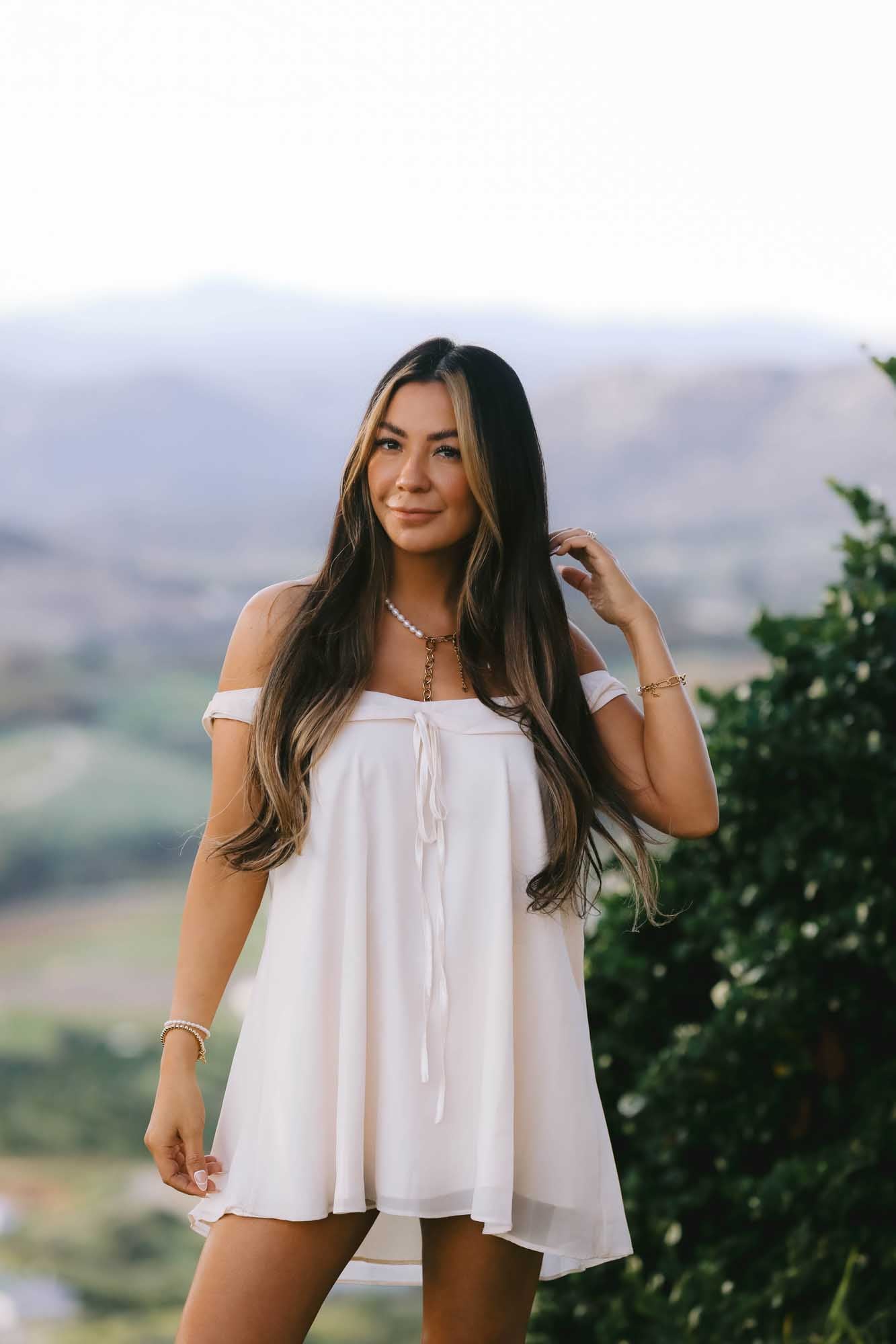 Woman wearing a white dress with multiple gold and pearl bracelets on each wrist, gold and pearl necklace.
