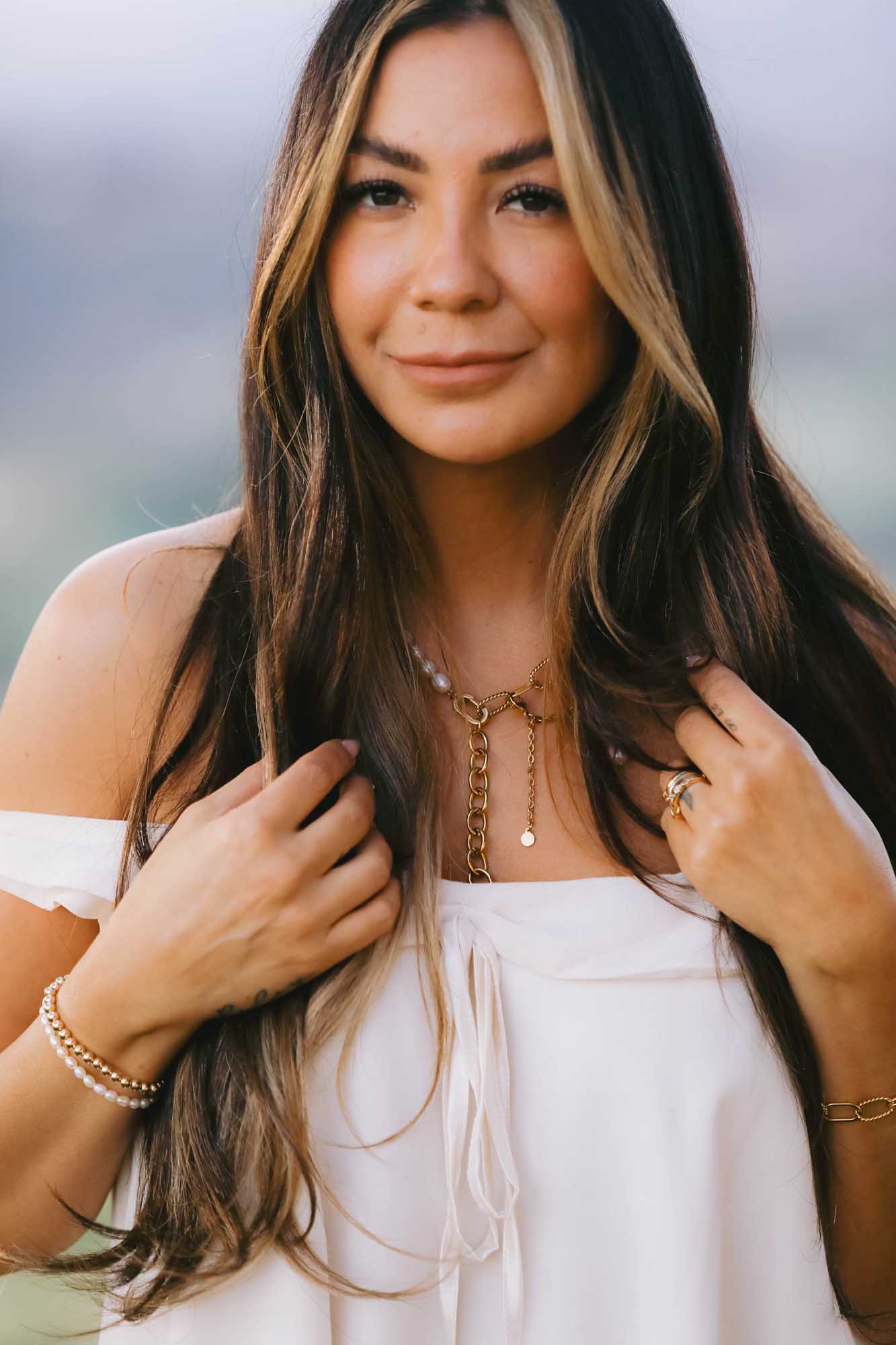 Woman with long hair wearing gold and pearl necklace, multiple gold and pearl bracelet, and gold ring.