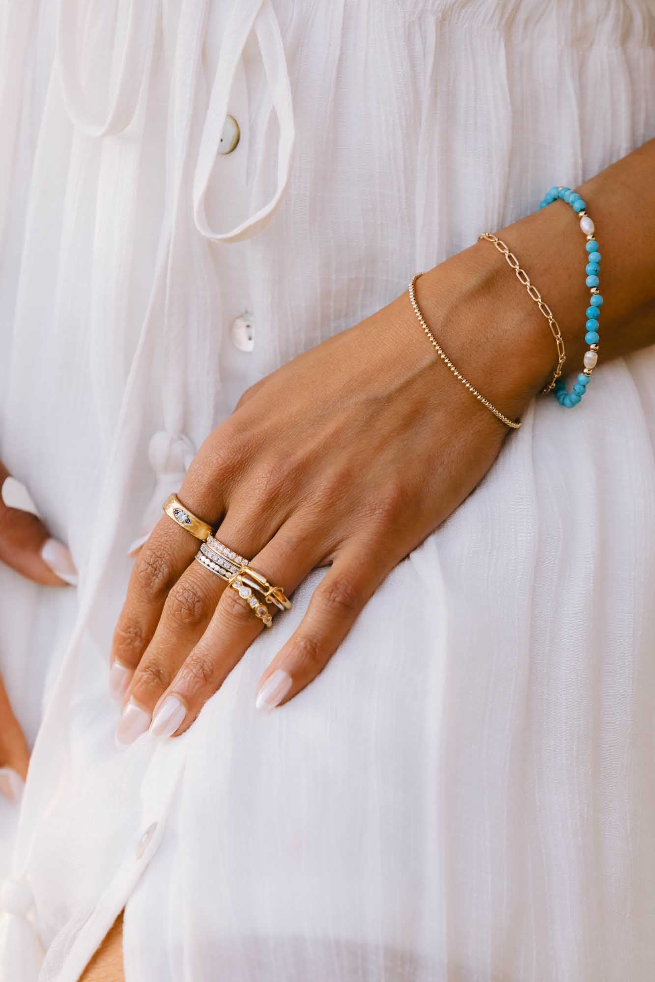 Close-up of a hand wearing multiple rings made of gold and silver along with two gold bracelets on a white background