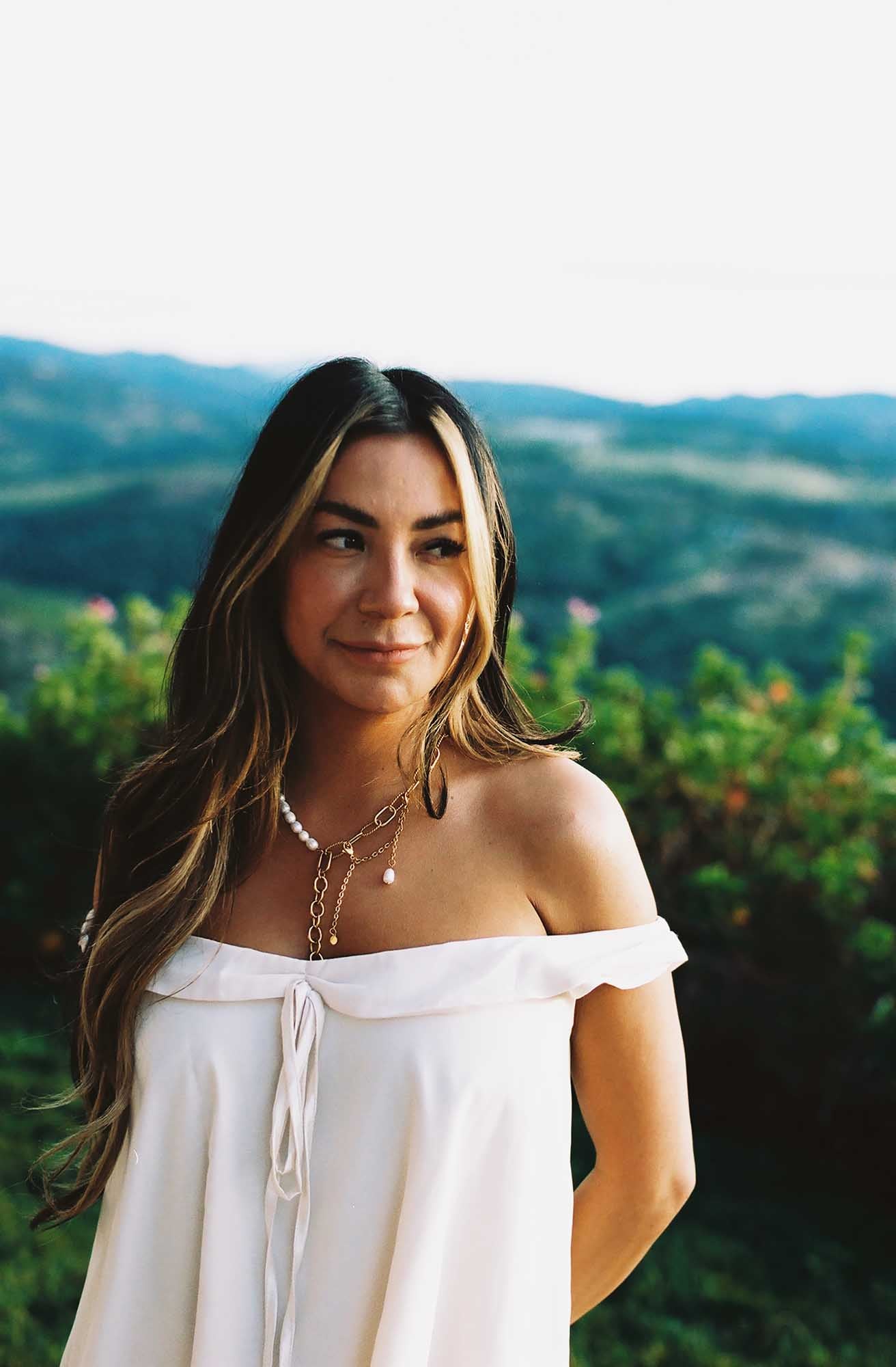 Woman in a white dress wearing a gold and pearl necklace on a blurred scenic landscape as a background.