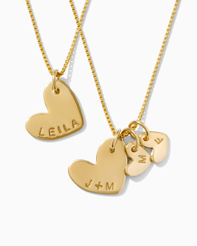 A gold heart necklace imprinted with the word 'Leila' and another gold necklace with three hearts imprinted with the letters 'J + M', 'M', and 'F' on a white background.