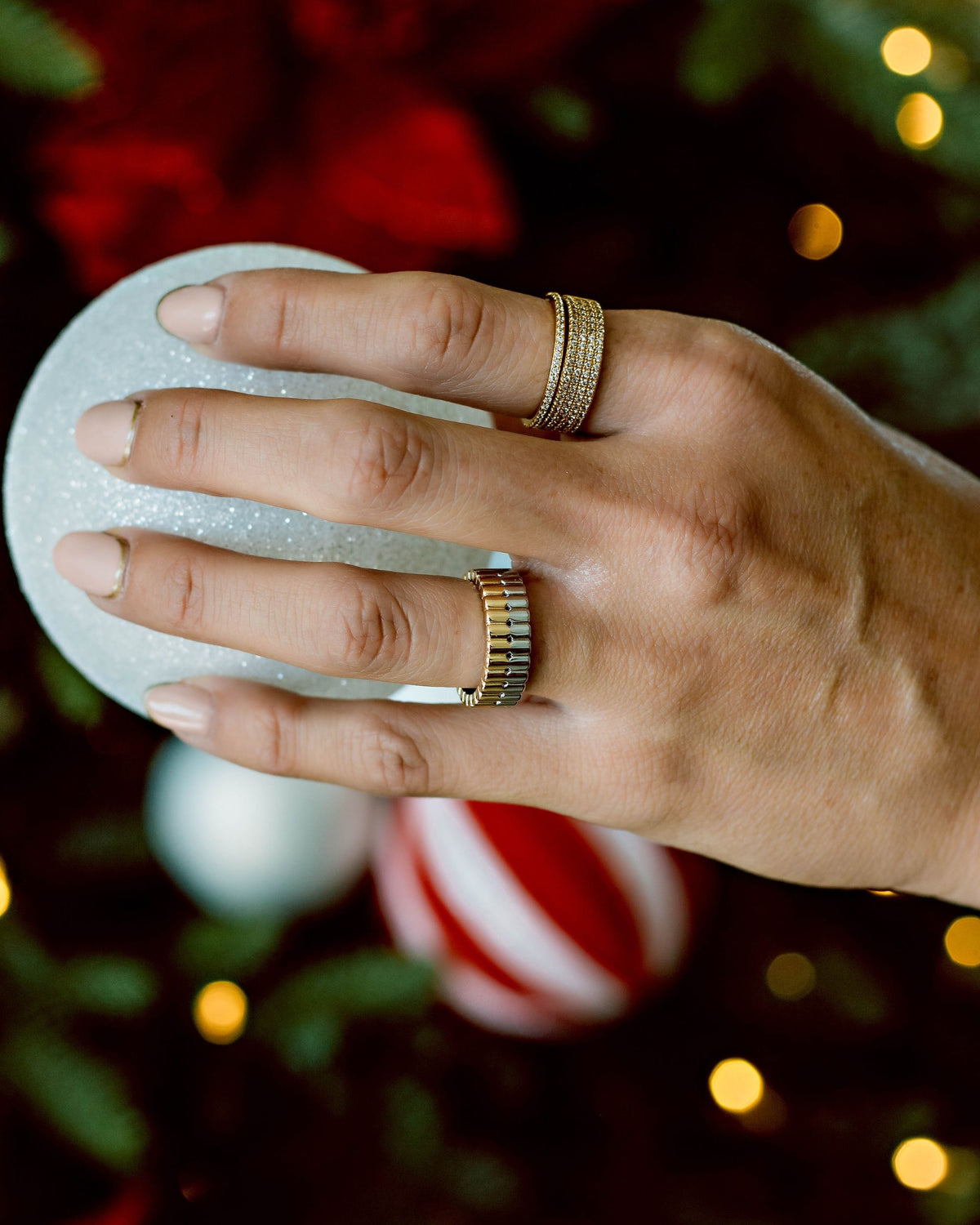 A close-up of a hand holding a Christmas ball decoration wearing a gold and silver ring along with two gold ring embedded with multiple cubic zirconia gemstone on a green blurred background.
